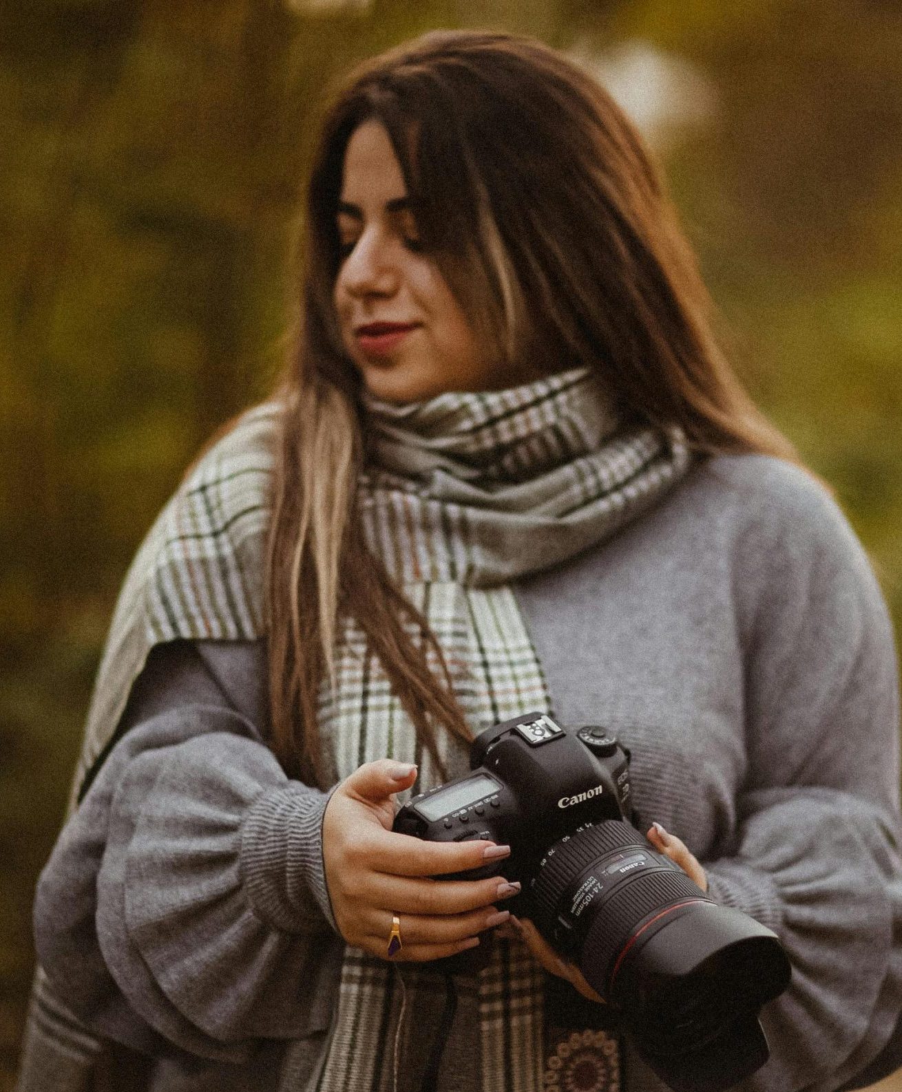 Middle Eastern woman outdoors holding a DSLR camera, wearing a scarf and gray sweater.