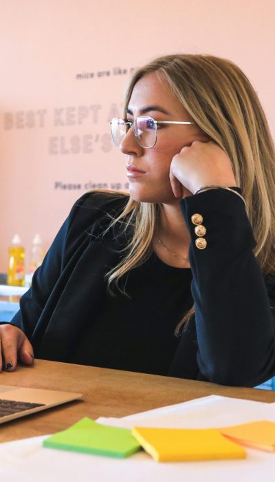 Blond woman in black blazer with glasses, working on laptop in modern office.